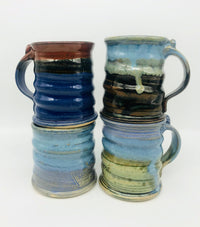 Four handmade pottery mugs are neatly stacked upon one another against a white background. From left: a brown and dark blue glaze, a light blue glaze, a light blue and brown glaze, and a light blue and light green glaze.